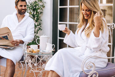 Happy couple sitting outside a hotel in white morning robes. Married couple smiling while having tea together in the morning. Couple enjoying a romantic holiday at a luxury resort. - JLPPF00938