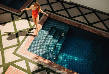 Aerial view of a woman standing next to a pool in red swimwear. Tourist woman feeling the swimming pool water with her foot. Woman vacationing at a luxury holiday resort in the summer. - JLPPF00952