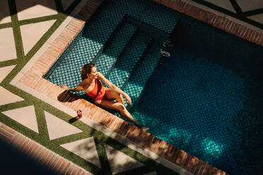 Glückliche Frau lächelt fröhlich, während sie sich in einem roten Badeanzug am Rande eines Swimmingpools entspannt. Frau genießt ihren Sommerurlaub in einem luxuriösen Urlaubsort. - JLPPF00966