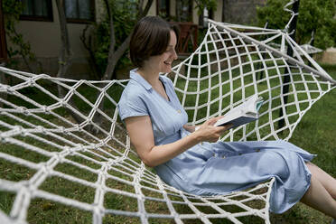 Lächelnde Frau, die auf einer Hängematte im Garten sitzt und ein Buch liest - ANNF00010