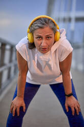 Mature woman wearing wireless headphones taking break from run at bridge - ZEDF04792