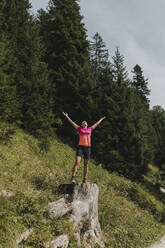 Hiker with arms outstretched standing on tree stump in forest - DMGF00835