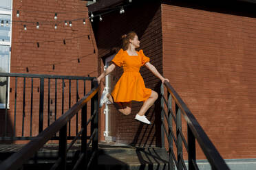 Frau in orangefarbenem Kleid balanciert auf dem Geländer einer Treppe - LLUF00842