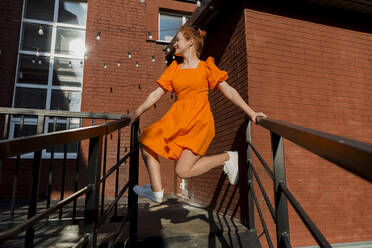 Lächelnde Frau in orangefarbenem Kleid balanciert auf einem Treppengeländer - LLUF00845