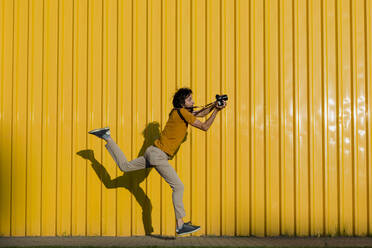 Mann, der durch die Kamera fotografiert, während er auf dem Fußweg vor einer gelben Wand läuft - LLUF00876