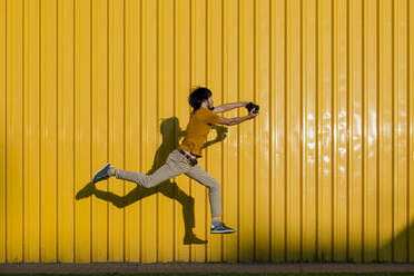 Mann fotografiert durch die Kamera und springt auf den Fußweg vor einer gelben Wand - LLUF00877