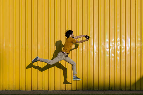 Man photographing through camera and jumping on footpath in front of yellow wall - LLUF00877