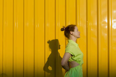 Woman wearing green dress standing with eyes closed in front of yellow wall - LLUF00878