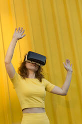 Woman wearing virtual reality simulator gesturing while standing in front of yellow wall - LLUF00887