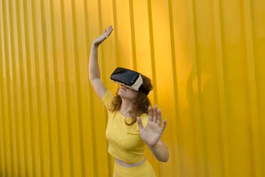 Smiling woman wearing virtual reality simulator gesturing in front of yellow wall - LLUF00889