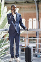 Lächelnder Geschäftsmann mit Gepäck, der am Geländer eines Bahnhofs mit seinem Smartphone telefoniert - IFRF01747