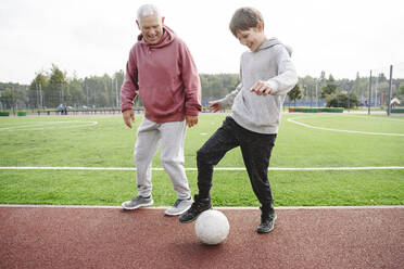 Glücklicher Junge mit Großvater spielt mit Fußball auf einem Sportplatz - EYAF02170
