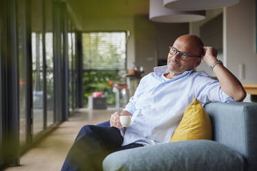 Nachdenklicher Mann mit Kaffeetasse, der zu Hause auf dem Sofa sitzt - RBF08900