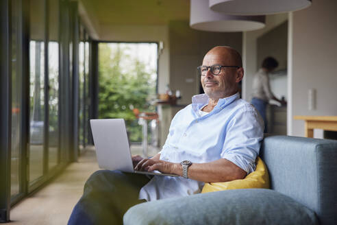 Nachdenklicher Mann mit Laptop, der zu Hause auf dem Sofa sitzt - RBF08903