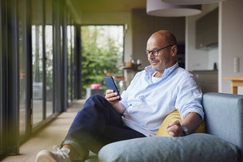 Senior man using smart phone sitting on sofa at home - RBF08904