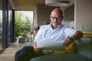Senior man reading documents sitting on sofa at home - RBF08907