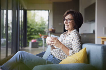 Happy senior woman with coffee cup sitting on sofa at home - RBF08915