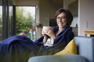 Happy woman with coffee cup and blanket sitting on sofa at home - RBF08917