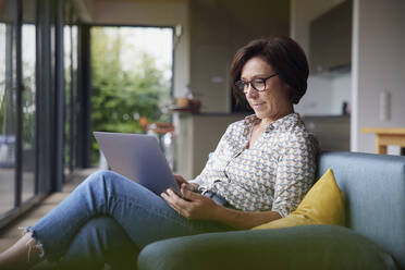 Woman using laptop sitting on sofa at home - RBF08922