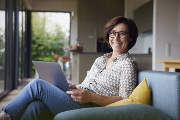 Glückliche Frau mit Laptop, die zu Hause auf dem Sofa sitzt - RBF08923