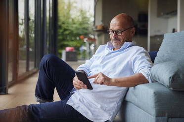 Senior man using smart phone leaning on sofa at home - RBF08925