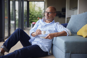 Thoughtful senior man with coffee cup sitting by sofa at home - RBF08927