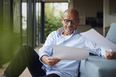 Senior man with documents sitting by sofa at home - RBF08928