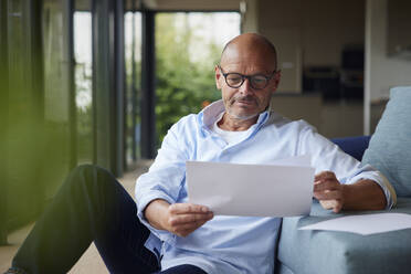 Senior man reading documents sitting by sofa at home - RBF08929