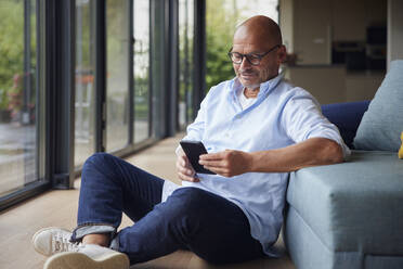 Smiling man using smart phone sitting on floor by sofa at home - RBF08931