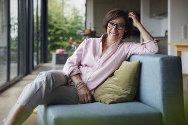 Happy woman with hand in hair sitting on sofa at home - RBF08943