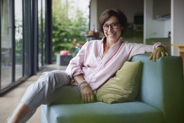 Happy senior woman wearing eyeglasses sitting on sofa at home - RBF08944