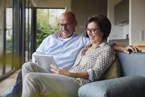 Glückliche ältere Frau mit Mann, der einen Tablet-PC auf dem Sofa zu Hause benutzt - RBF08954