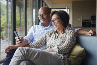 Glückliche Frau mit Mann, der ein Smartphone benutzt, sitzt auf dem Sofa zu Hause - RBF08958