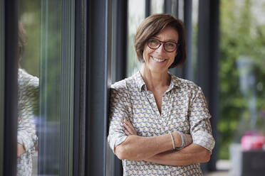 Happy senior woman with arms crossed leaning on window - RBF08967