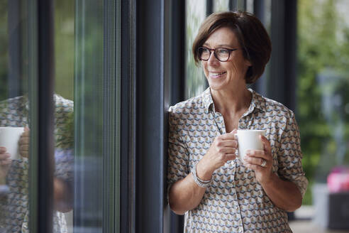 Happy senior woman with coffee cup leaning on window at home - RBF08969