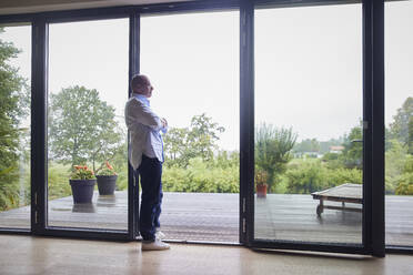 Nachdenklicher älterer Mann mit verschränkten Armen, der sich zu Hause an eine Glastür lehnt - RBF08973