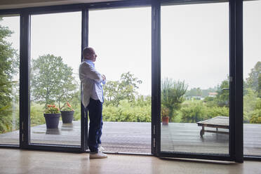 Thoughtful senior man with arms crossed leaning on glass door at home - RBF08973