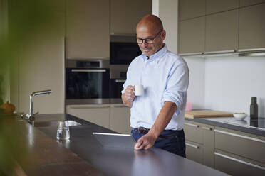 Lächelnder Mann mit Kaffeetasse und Tablet-PC an der Kücheninsel stehend - RBF08985