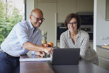 Glücklicher Mann mit Frau, die einen Laptop auf dem Küchentisch zu Hause benutzt - RBF08992