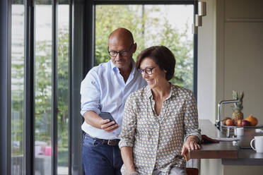 Lächelnde Frau mit Mann, der sein Smartphone an der Kücheninsel zu Hause benutzt - RBF09001