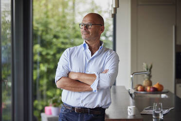Thoughtful senior man with arms crossed standing by kitchen island at home - RBF09015