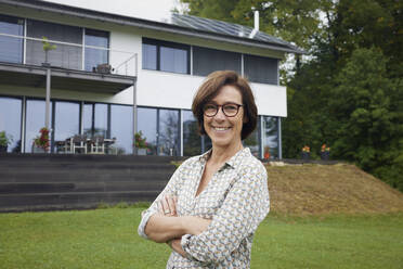 Happy senior woman with arms crossed standing in front of house - RBF09023