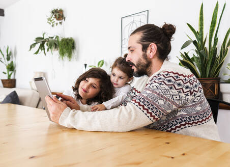 Glücklicher Vater mit Bart im warmen Pullover sitzt mit Kindern am Tisch und benutzt ein Tablet in der Wohnung - ADSF38823