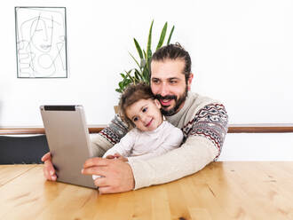 Bearded male in sweater sitting at table with daughter using tablet in light apartment with potted plants - ADSF38824