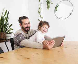 Bärtiger Mann im Pullover sitzt mit Tochter am Tisch und benutzt ein Tablet in einer hellen Wohnung mit Topfpflanzen - ADSF38826