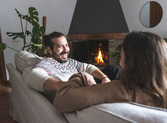 Smiling male and unrecognizable female sitting together on sofa holding hands while laughing and looking at each other in apartment with fireplace - ADSF38829