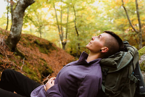 Junger Tourist in Freizeitkleidung mit Rucksack, der auf einem Baum liegt und eine Pause in einem herbstlichen Wald macht - ADSF38833