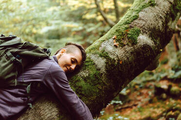 Junger Tourist mit geschlossenen Augen in Freizeitkleidung mit Rucksack auf einem Baum liegend und in einem herbstlichen Wald reisend - ADSF38839