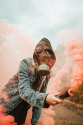 Person mit Gasmaske und Haube hält Bombe mit rosa Rauch in der erhobenen Hand in der Landschaft - ADSF38841
