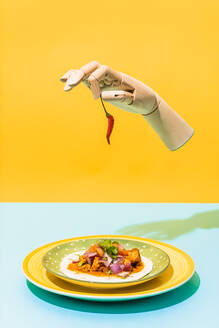 Künstliche Hand mit roter Chilischote schwebt über Teller mit mexikanischem Hühnersalat auf blauem und gelbem Hintergrund - ADSF38849